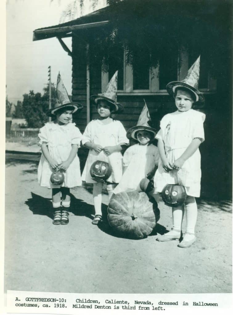 Four children in white dresses and pointed hats hold pumpkin buckets, with a large pumpkin on the ground.