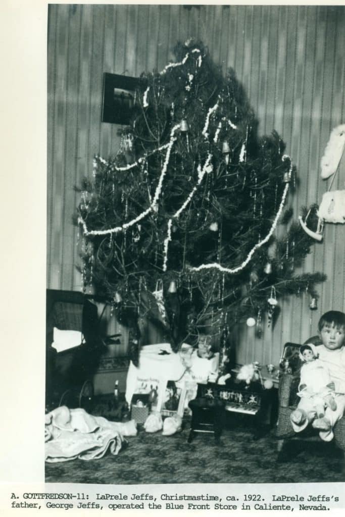 A black and white photo shows a decorated Christmas tree with a child holding a doll in a wicker chair.