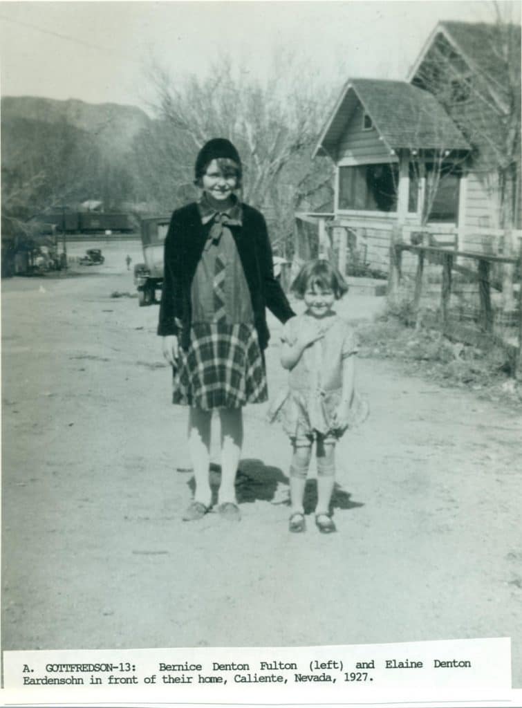 Two young girls, one older in a plaid dress and one younger in a light dress, stand on a dirt road.