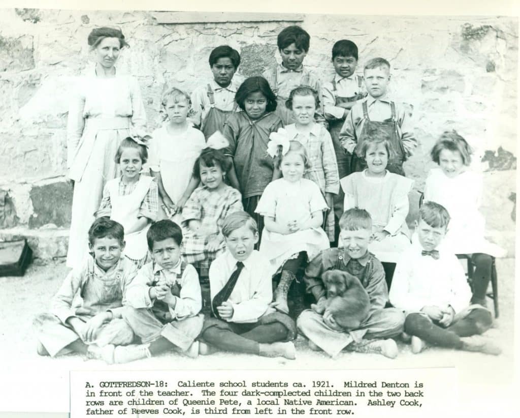 A teacher and 17 students, mostly children, pose in front of a stone wall in 1921.