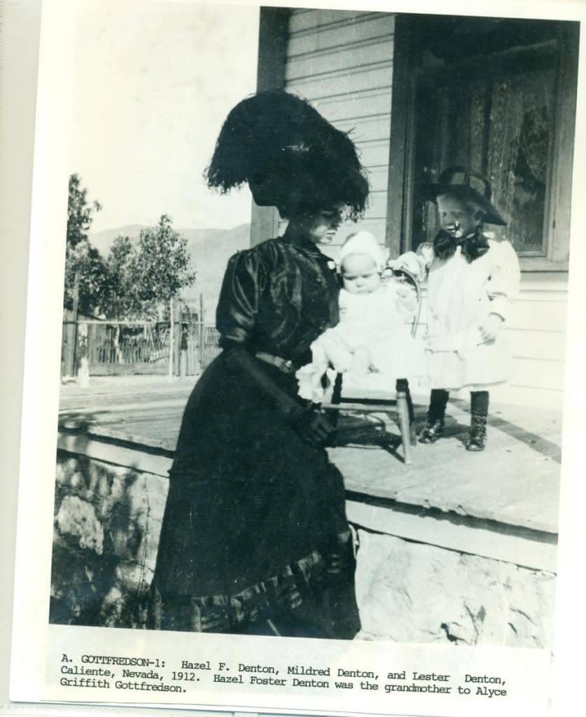 A woman in a feathered hat holds a baby, while a child in a hat stands nearby on a porch.