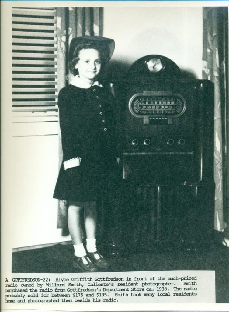 A young girl in a dark dress and hat stands smiling next to a large, dark, antique radio.