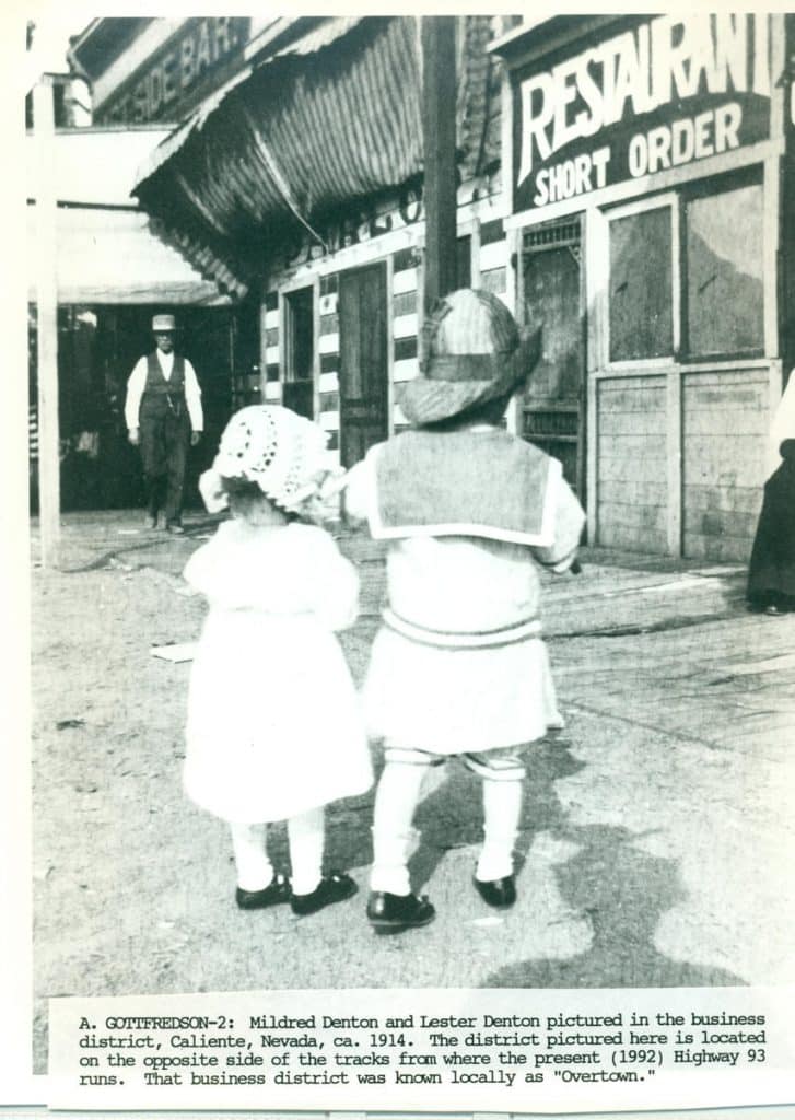 Two children, seen from behind, stand on a dirt path facing a "RESTAURANT SHORT ORDER" building.