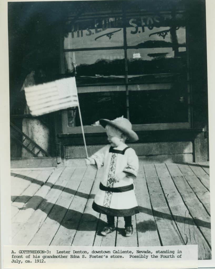 A young child in a hat and dress holds an American flag on a wooden porch in front of a store window.