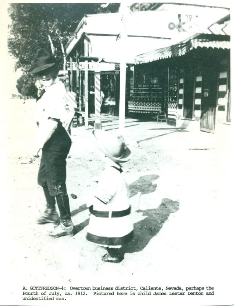 A man in a hat and a child in a white outfit stand on a dirt street in front of a building.