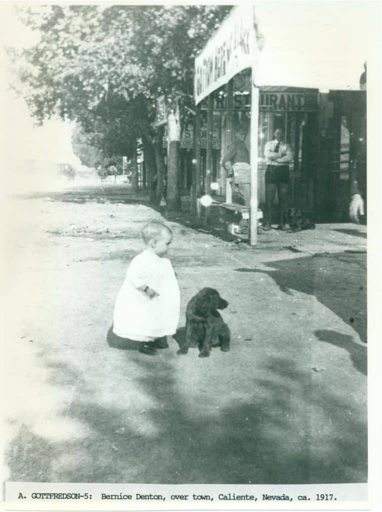 A baby in a white dress sits next to a dark dog on a dirt street in front of a "RESTAURANT."