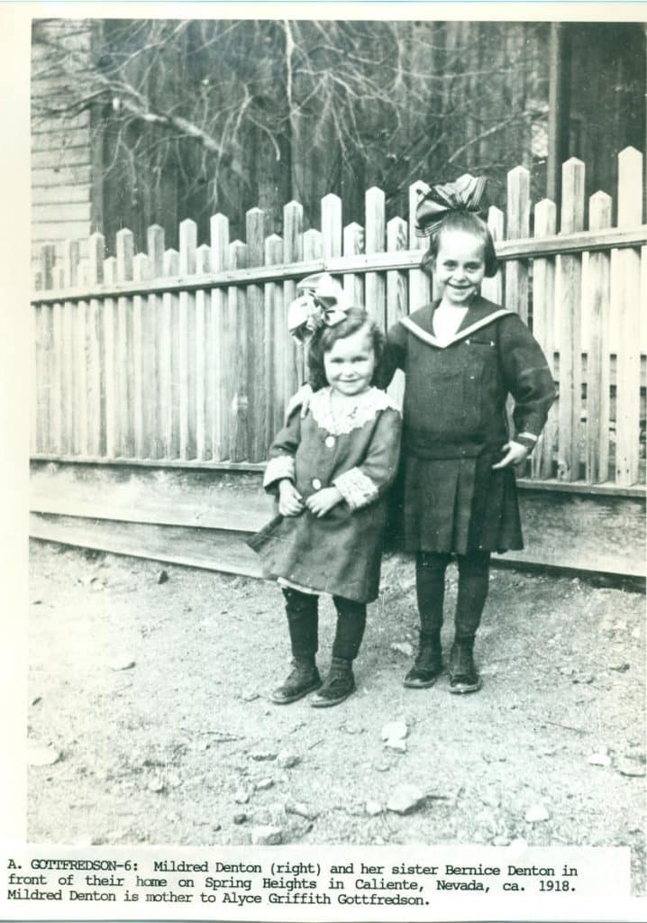 Two smiling young girls stand in front of a picket fence, one in a coat and the other in a sailor dress.