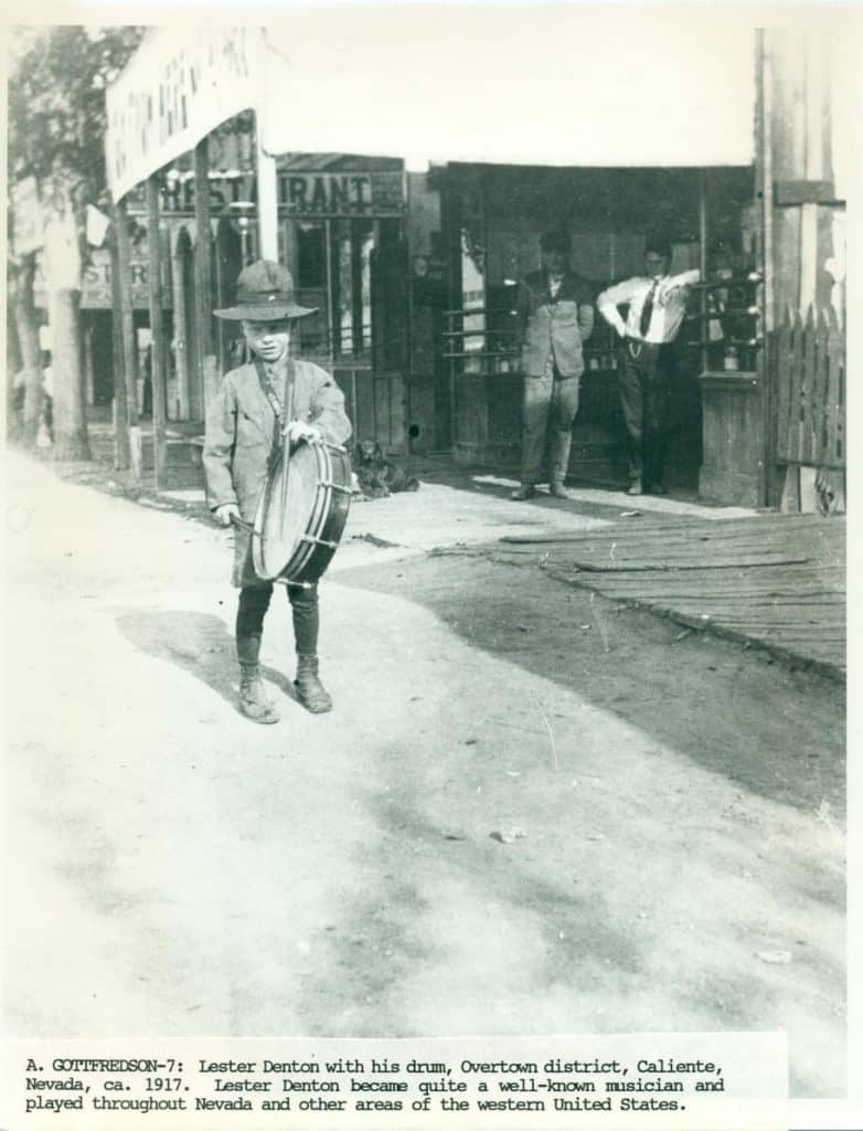 A young boy in a hat and suit holds a drum on a dirt road in front of a building with "RESTAURANT" visible.