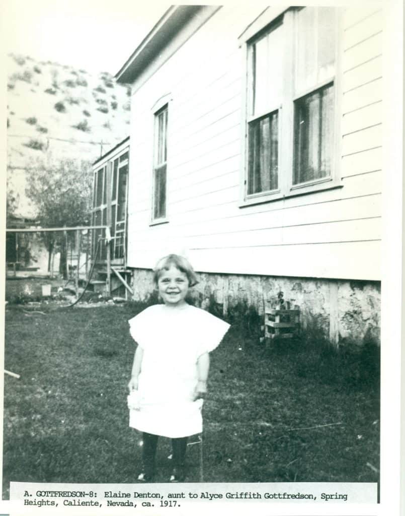 A smiling young girl in a white dress stands in a grassy yard in front of a white house with a screened porch.