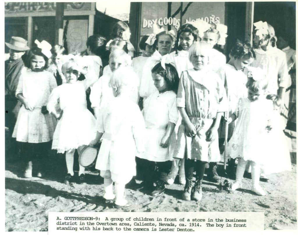 A group of children in white dresses and light-colored clothes stands in front of a "DRY GOODS" store.