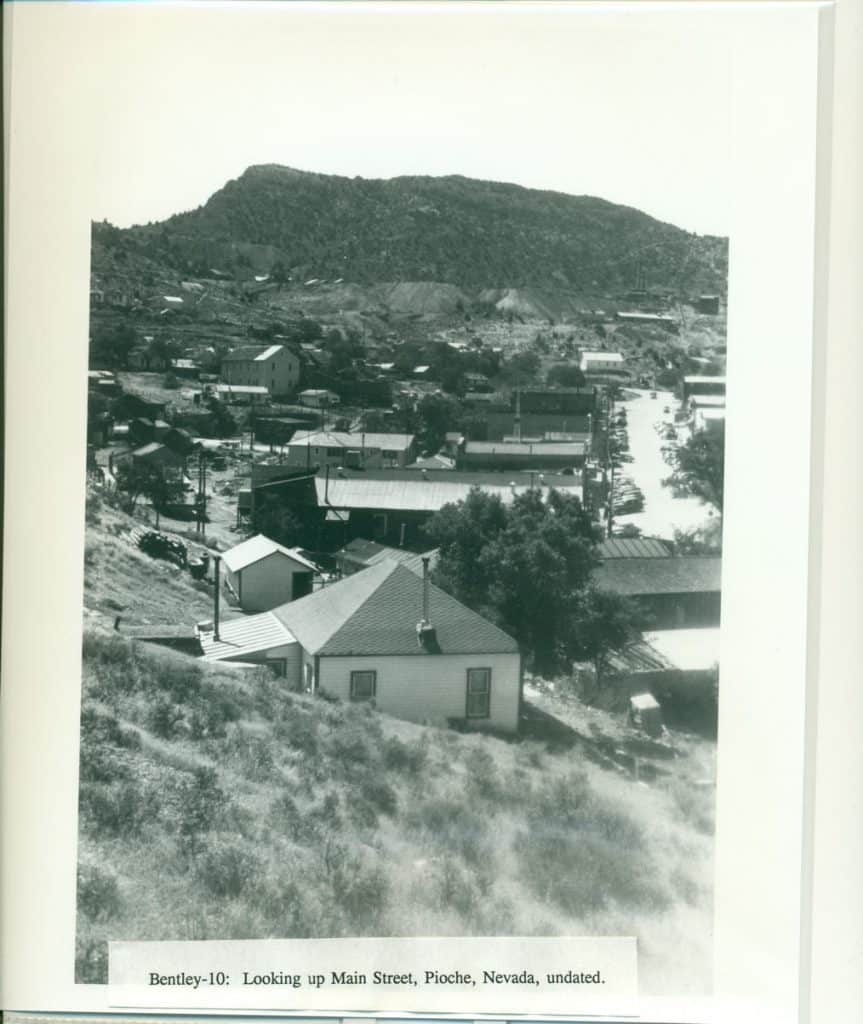 A black and white photo shows a town with buildings and a street, nestled below a large, treed mountain.