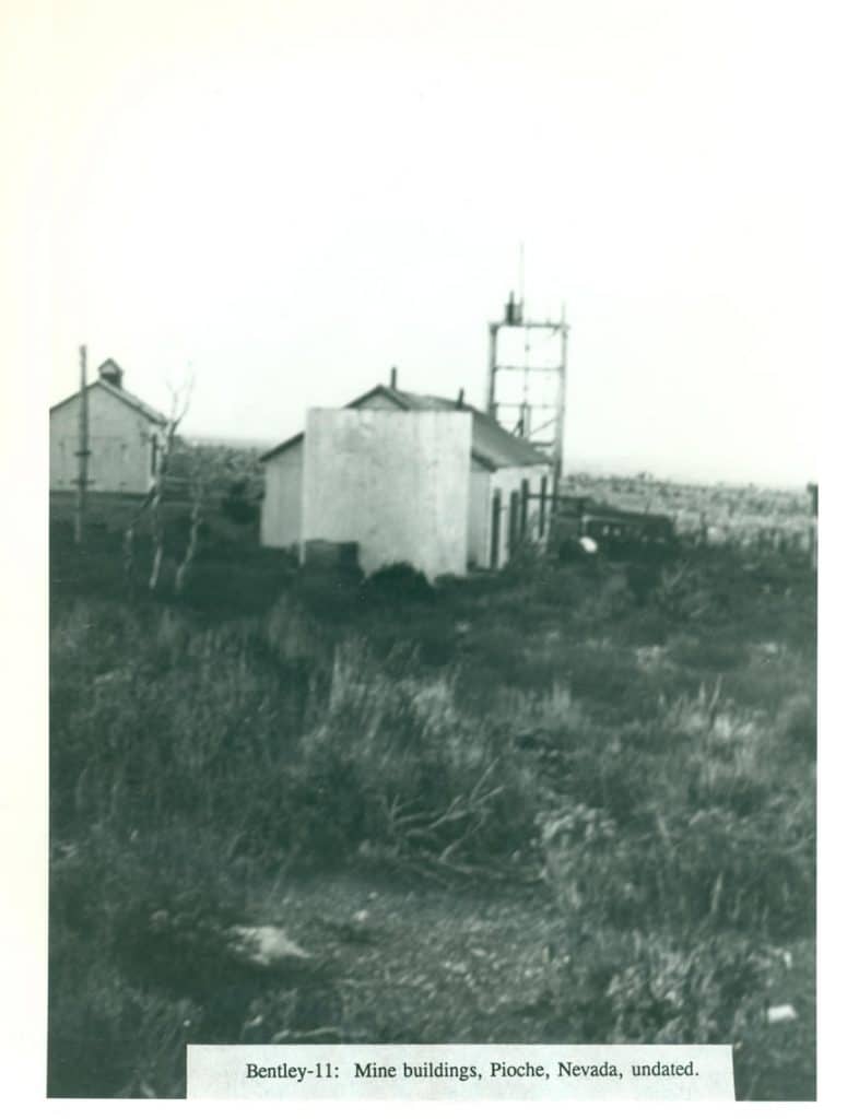 Black and white photo of three mine buildings and a metal tower on a grassy hill under a bright sky.