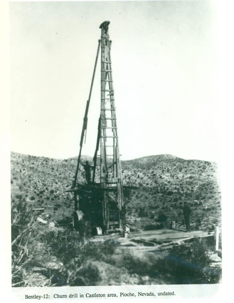 A tall, wooden churn drill stands in a desolate, hilly landscape with two men near its base.