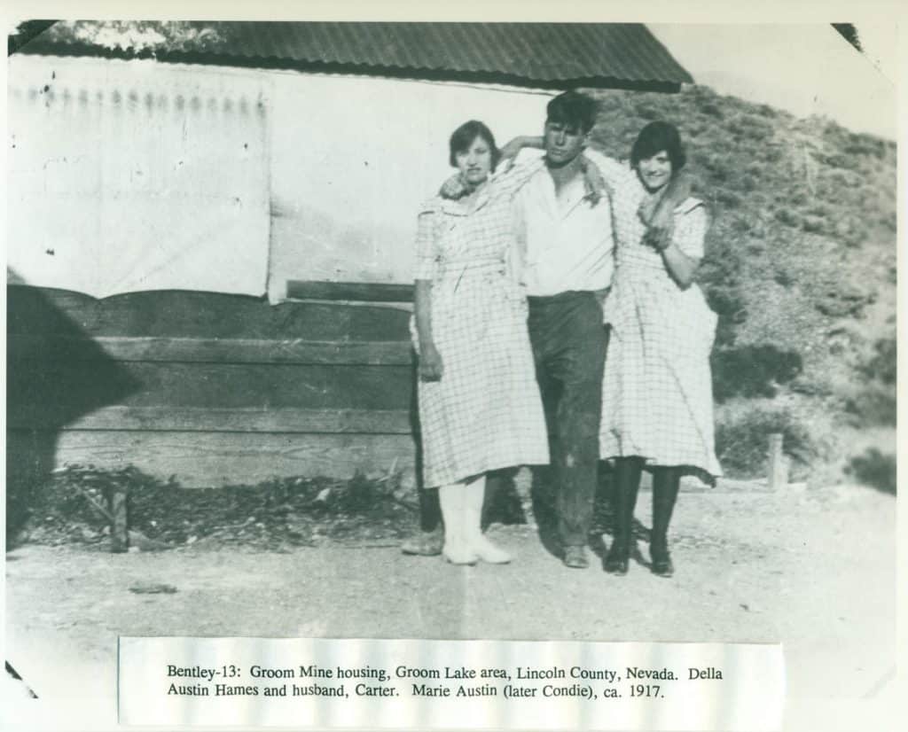 Three young adults, two women and a man, stand arm-in-arm in front of a wooden building and dirt hill.