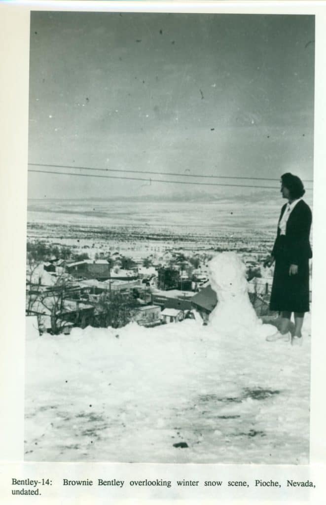 A woman in a dark coat and light shirt stands next to a snowman, overlooking a snow-covered town.