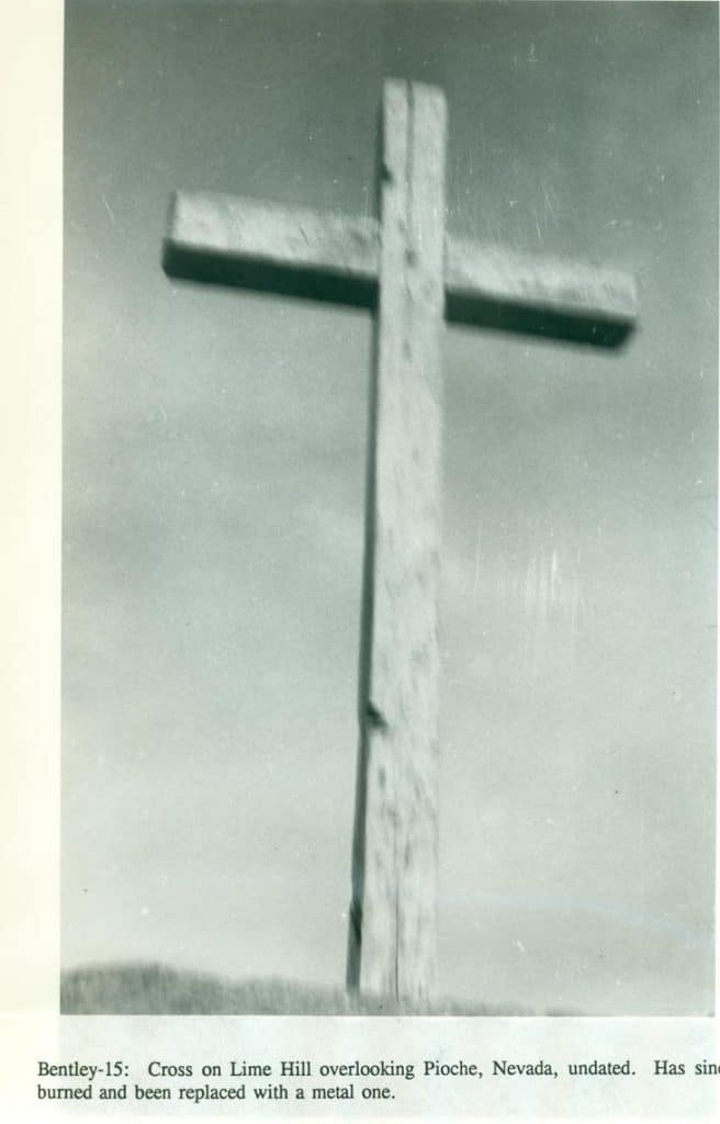 A weathered wooden cross stands against a light, hazy sky with "Bentley-15: Cross on Lime Hill overlooking Pioche, Nevada, undated. Has since burned and been replaced with a metal one."