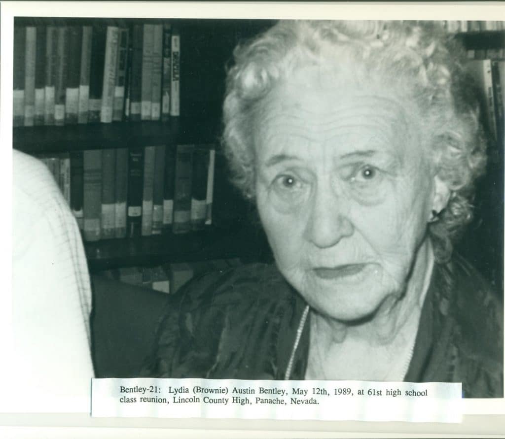 A black and white photo shows an older woman with curly white hair looking forward, with bookshelves behind her.