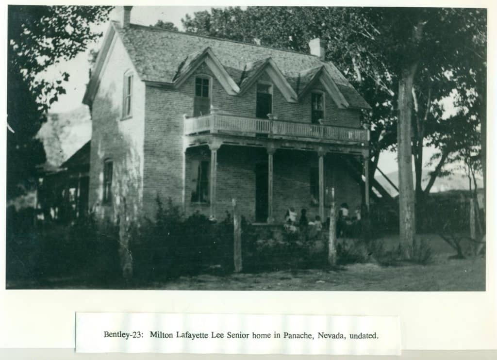 A two-story brick house with a second-floor balcony and three dormers is surrounded by trees and brush.