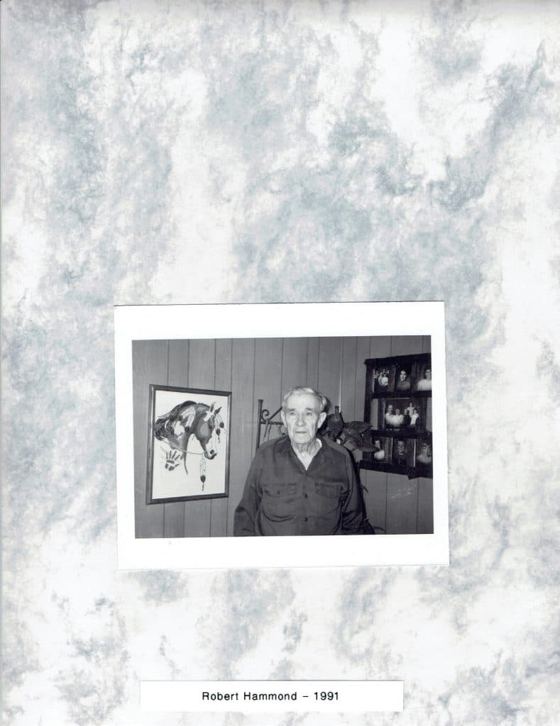 A black and white photo shows an older man in a dark shirt, with a horse painting and framed photos behind him.