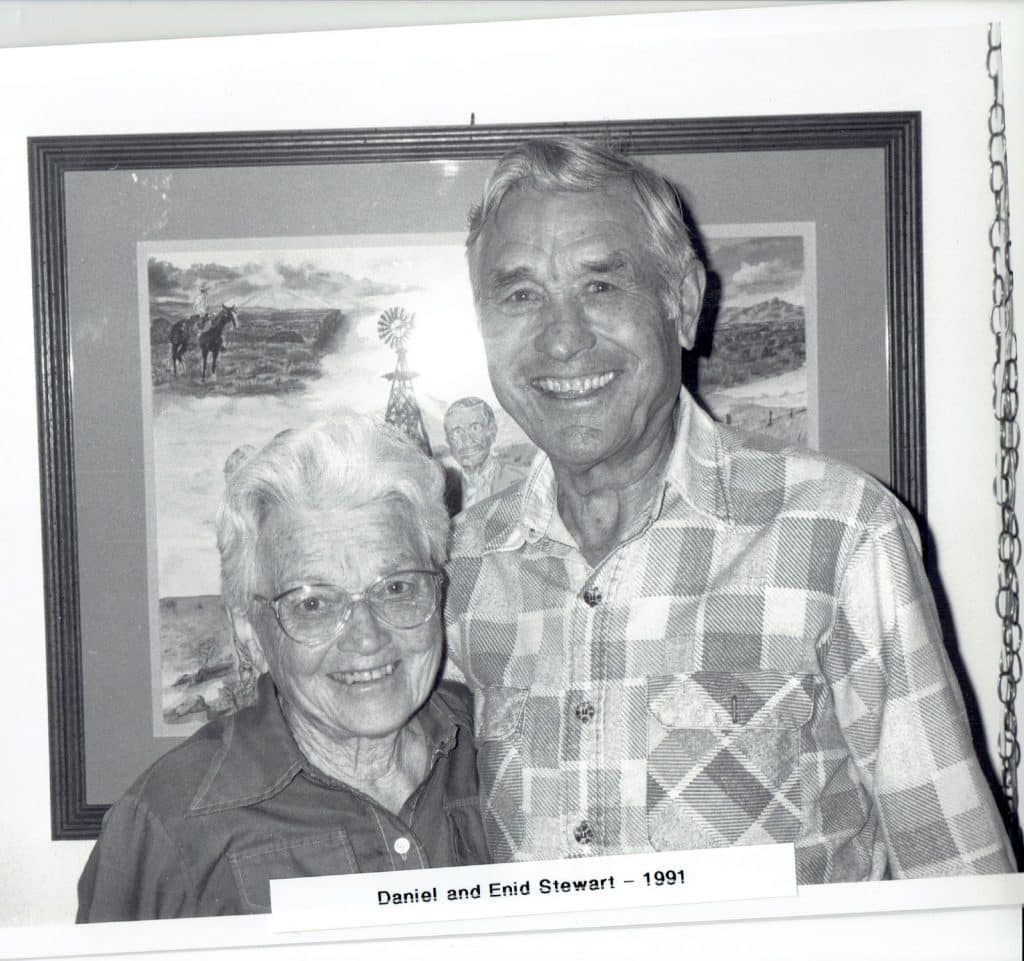 A smiling elderly woman in glasses and a man in a plaid shirt stand before a framed landscape.