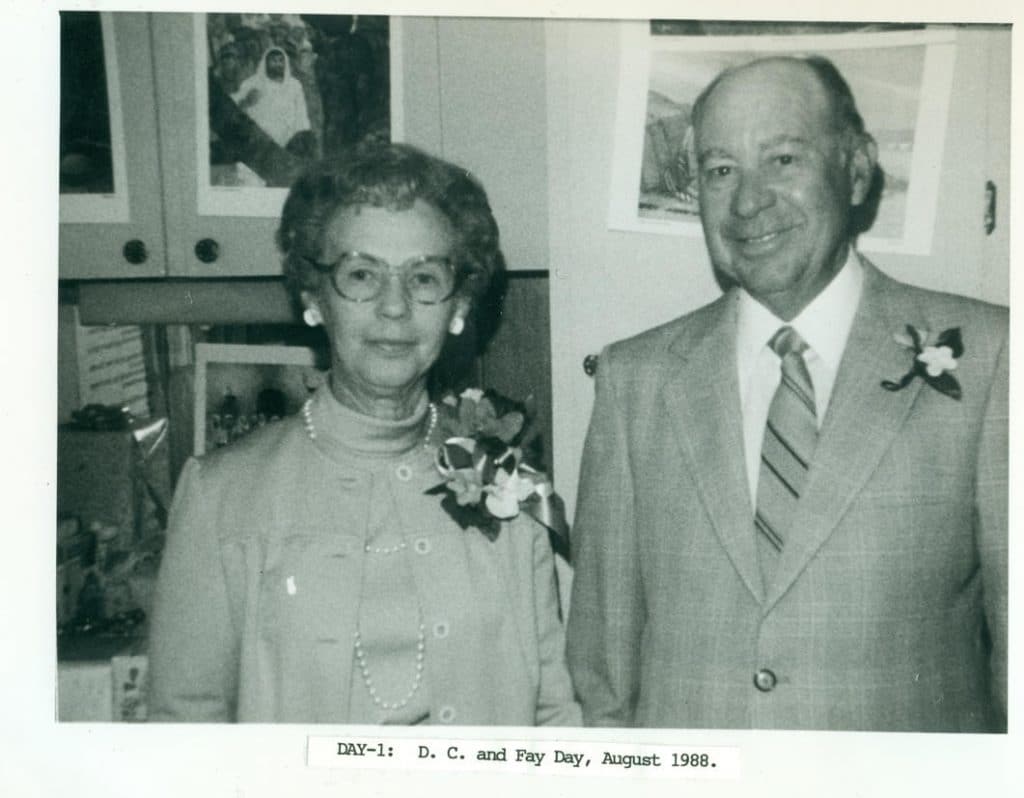 A smiling man and woman, both wearing corsages, stand in front of framed pictures. "DAY-1: D. C. and Fay Day, August 1988."