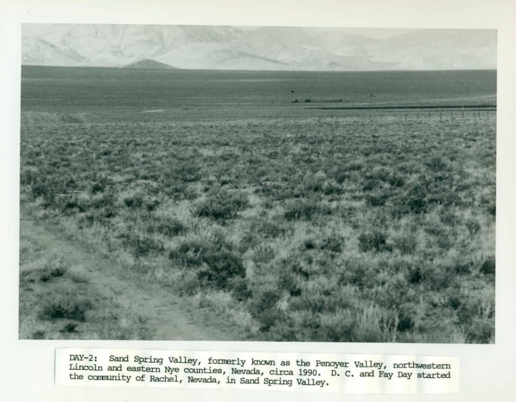 A black and white photo shows a vast, flat desert landscape covered in low-lying brush, with distant mountains.