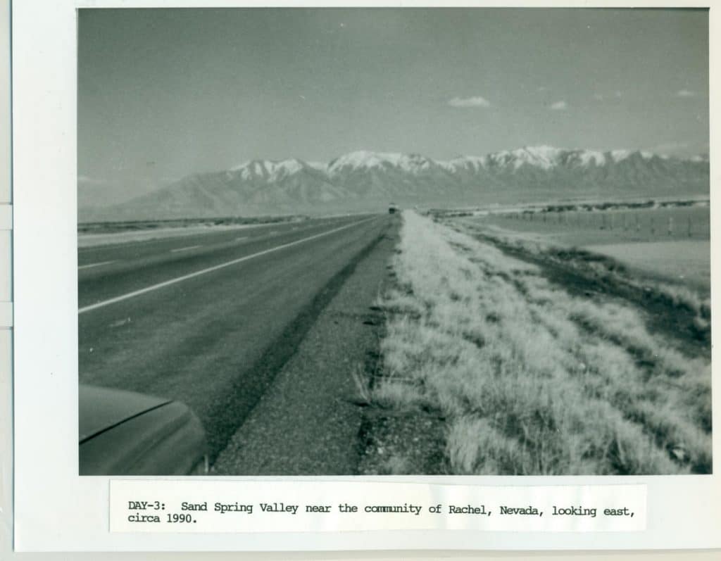 A black and white photo shows a long, straight road leading to distant snow-capped mountains.