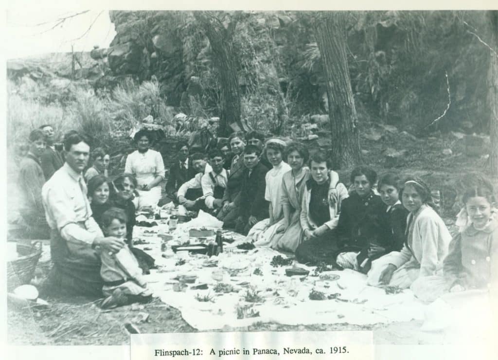 A black and white photo shows a large group of adults and children on a picnic blanket outdoors.
