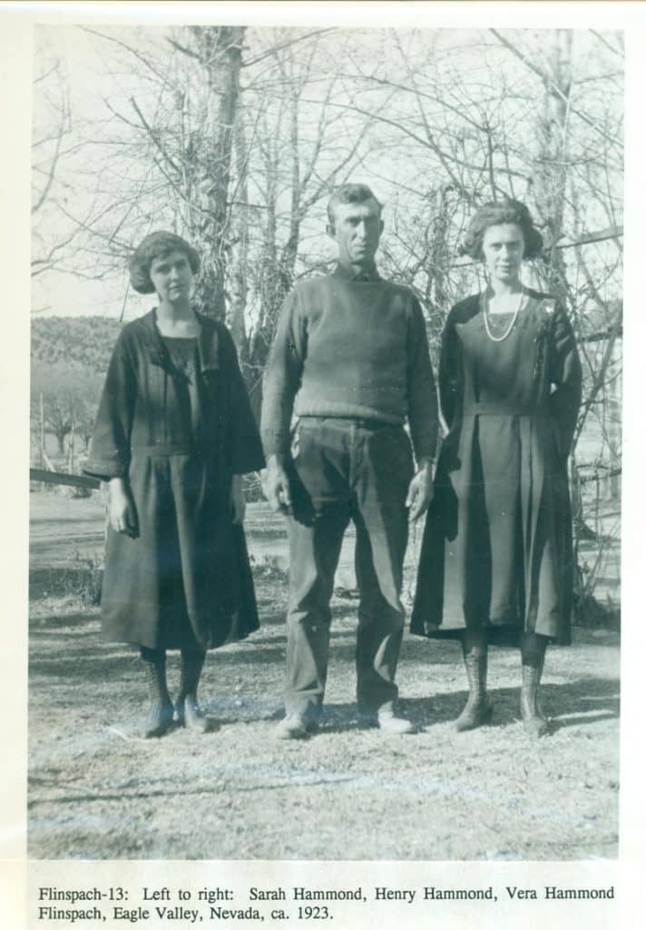 A man in a sweater and two women in dark dresses stand outdoors with bare trees behind them.