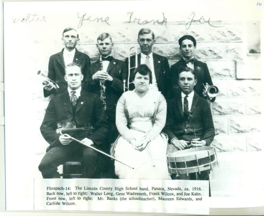 A black and white photo of seven band members with instruments in front of a stone wall.