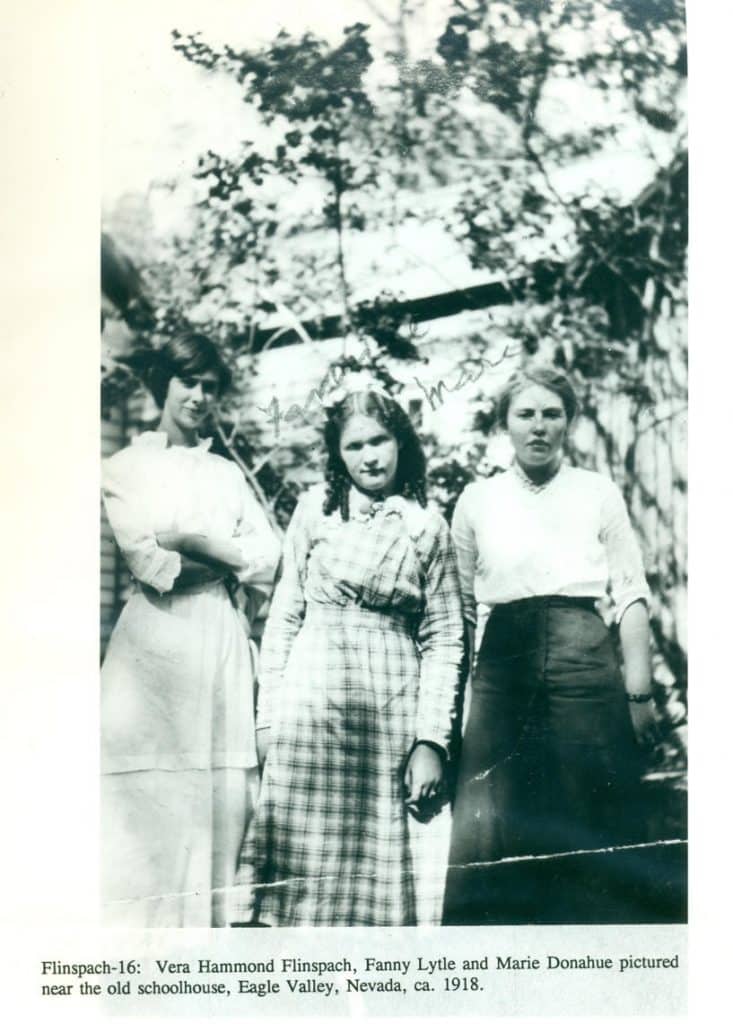 Three young women, two with dark hair, stand outdoors in long dresses with trees in the background.