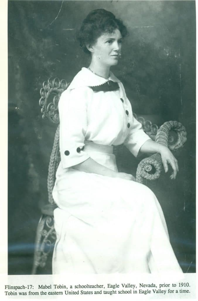 A young woman with curly hair, wearing a white dress with black buttons, sits in a wicker chair.