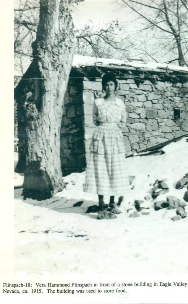 A young woman in a plaid dress stands in the snow next to a tree and a stone building.