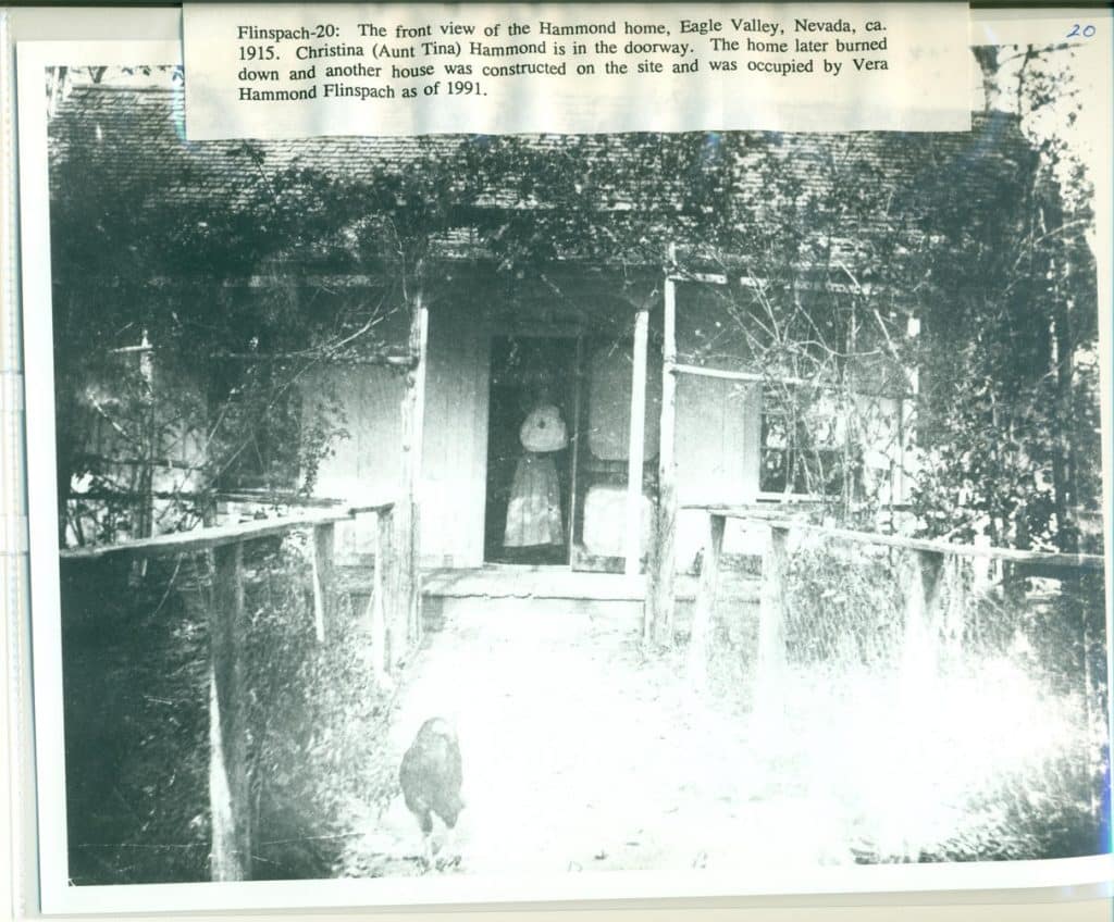 A grayscale photo shows a woman in a long dress and hat standing in the doorway of a house.