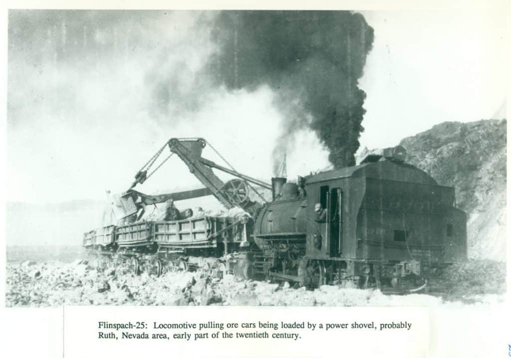 A black-and-white photo shows a steam locomotive pulling ore cars being loaded by a power shovel.