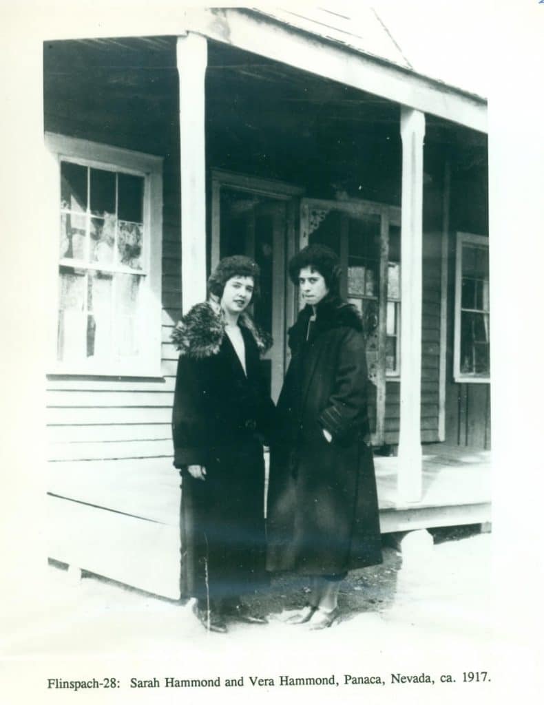 Two women in long, dark, fur-collared coats stand on a porch in front of a wooden house.