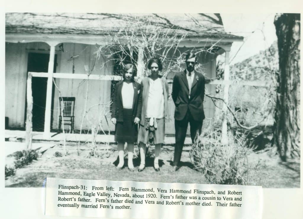 Three people stand in front of a house with a porch, surrounded by bare branches and dry grass.