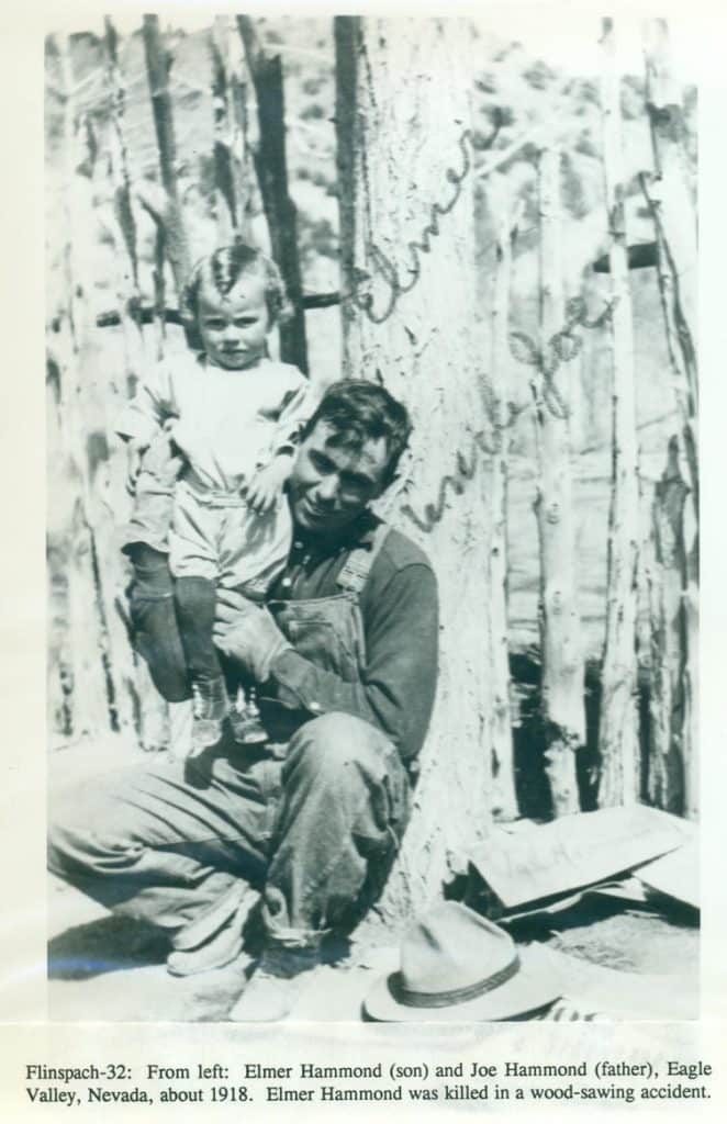 A smiling man in overalls holds a small child on his shoulders, standing before a wooden fence.