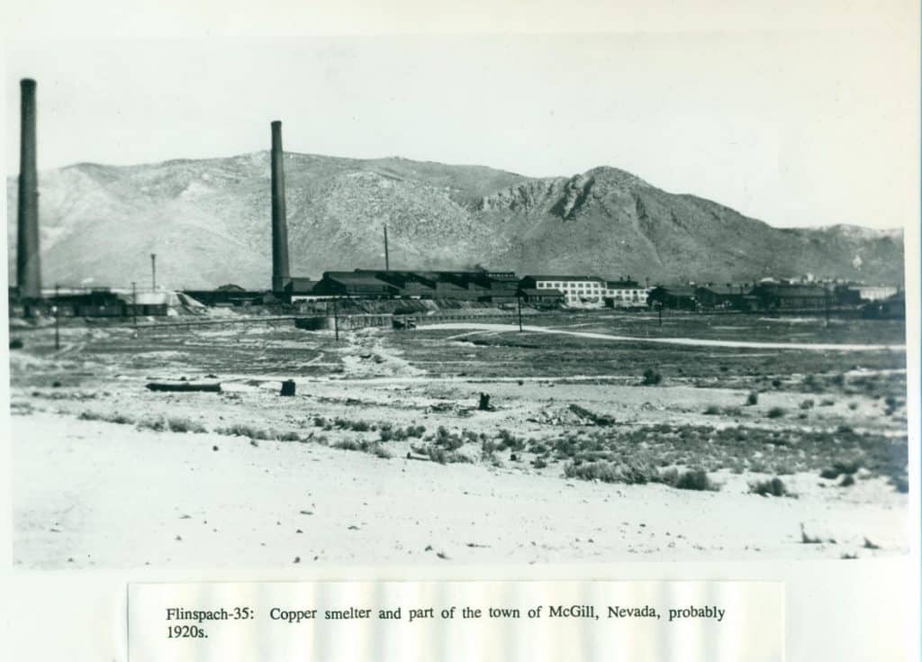 A black and white photo shows a copper smelter with two tall smokestacks and buildings in front of a mountain range.