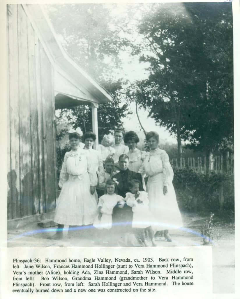 A black and white photo shows nine women and three children in white dresses standing outside a wooden house.