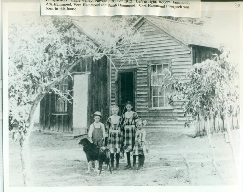 Four children and a dog stand in front of a wooden house with "Vera Hammond Flinspach was born in this house" visible at the top.