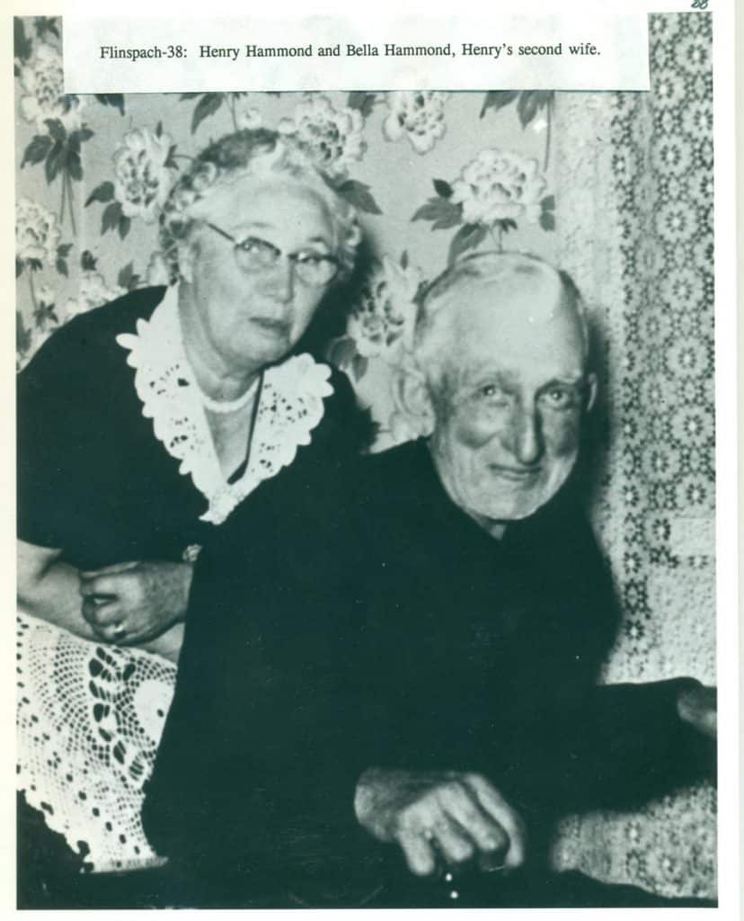 A smiling elderly man in a dark suit sits next to a woman with glasses and a white lace collar.