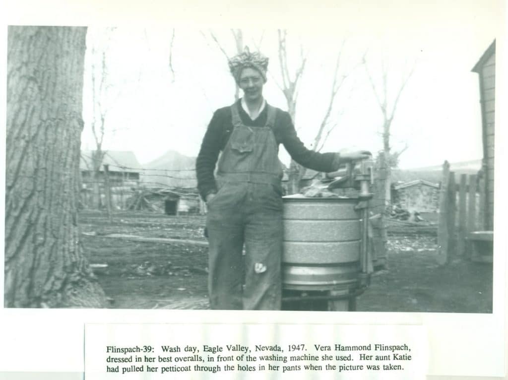 A woman in overalls and a headscarf stands next to a wringer washing machine outdoors.