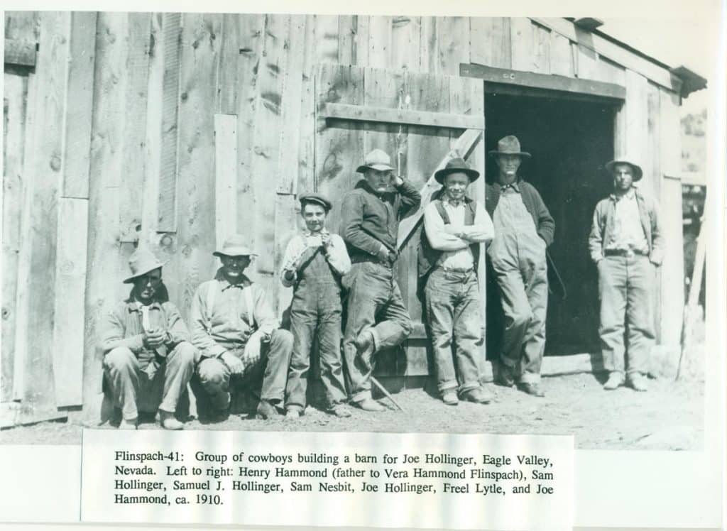 Seven men and a boy in cowboy hats and work clothes stand or sit in front of a wooden barn.