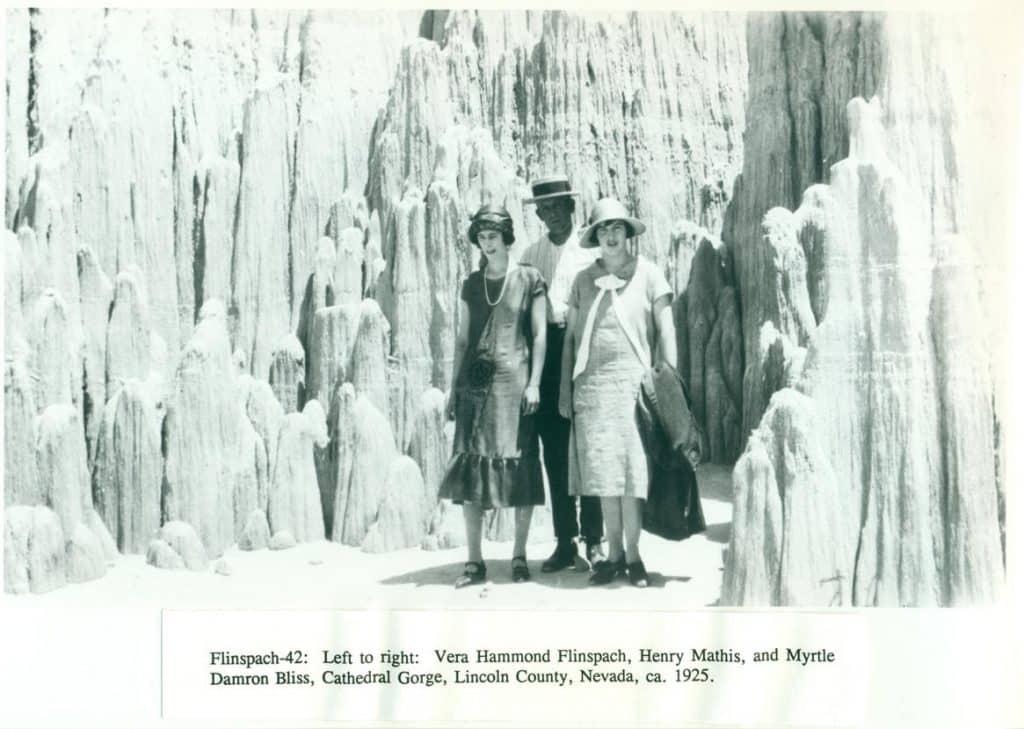 Three people, two women and one man, stand among tall, narrow rock formations under a bright sky.