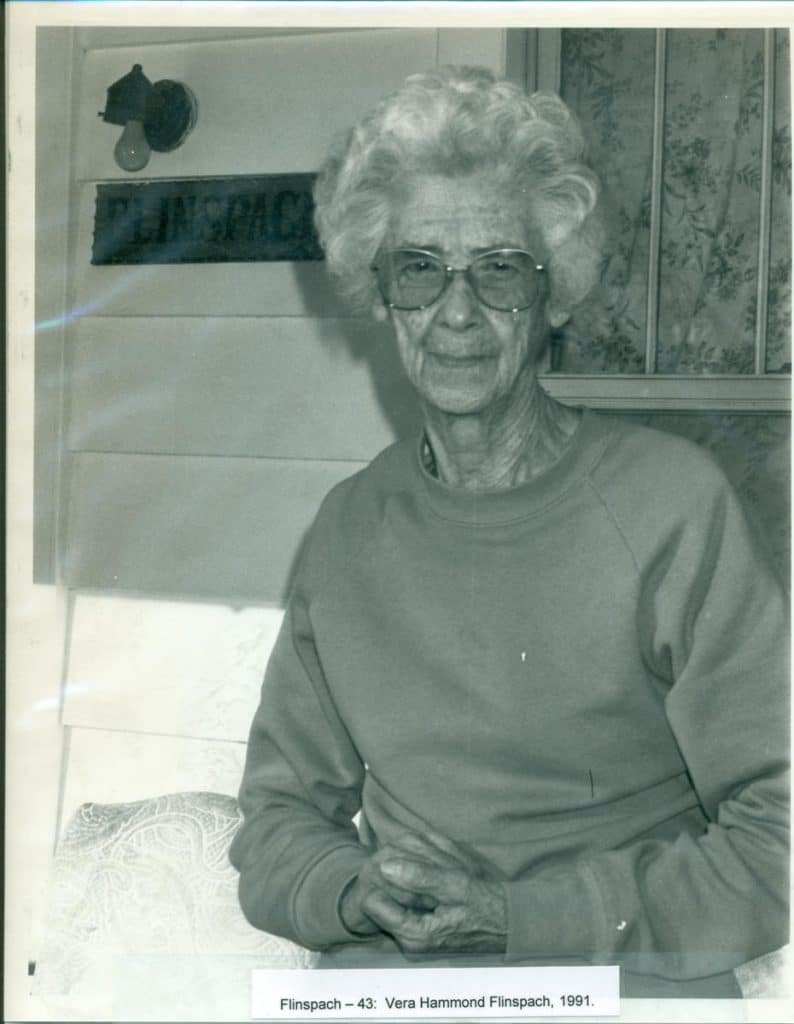 A smiling older woman with curly white hair and glasses wears a sweatshirt, with "FLINSPACH" on a sign behind her.