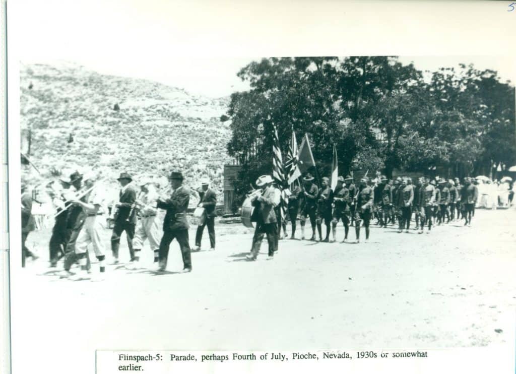 A black and white photo of a parade with a marching band, men carrying flags, and soldiers in uniform.