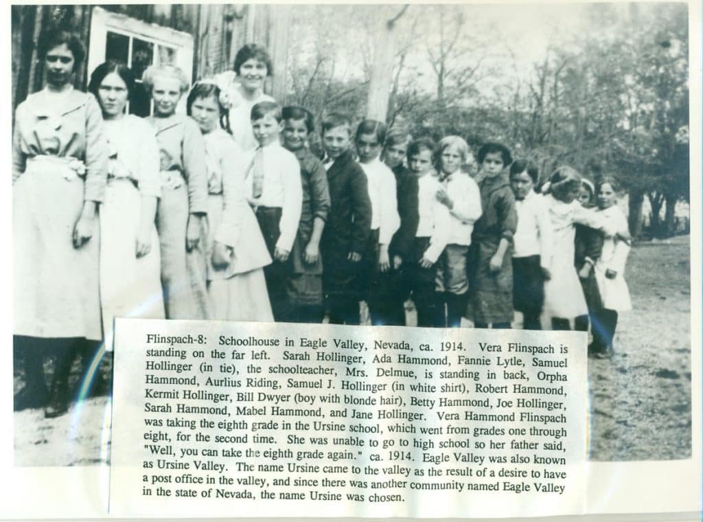 A group of 18 children and one adult stand in front of a building with trees in the background.