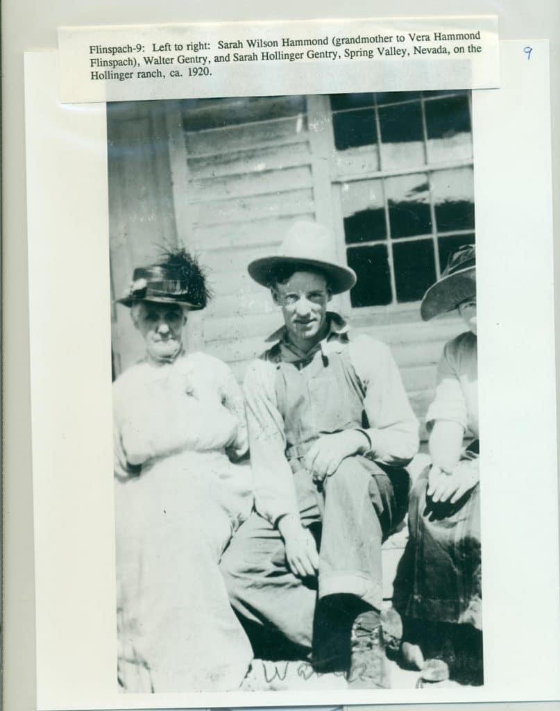 Three people, two women and a man in a hat and overalls, sit outside a building with a window.