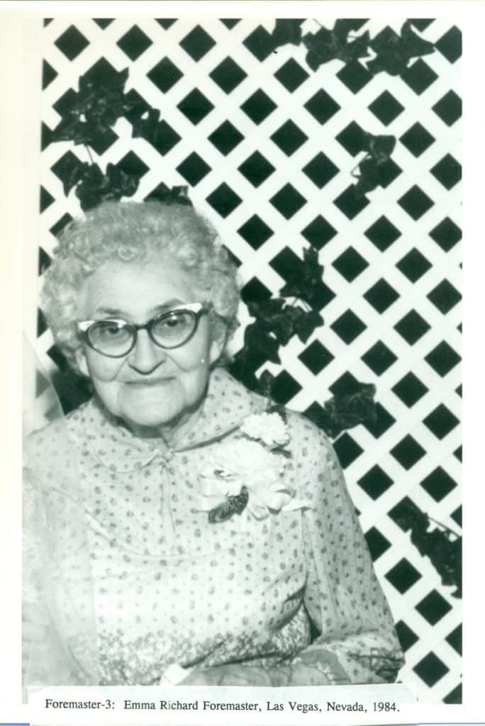 An elderly woman with curly white hair and cat-eye glasses smiles, wearing a patterned blouse with a flower.